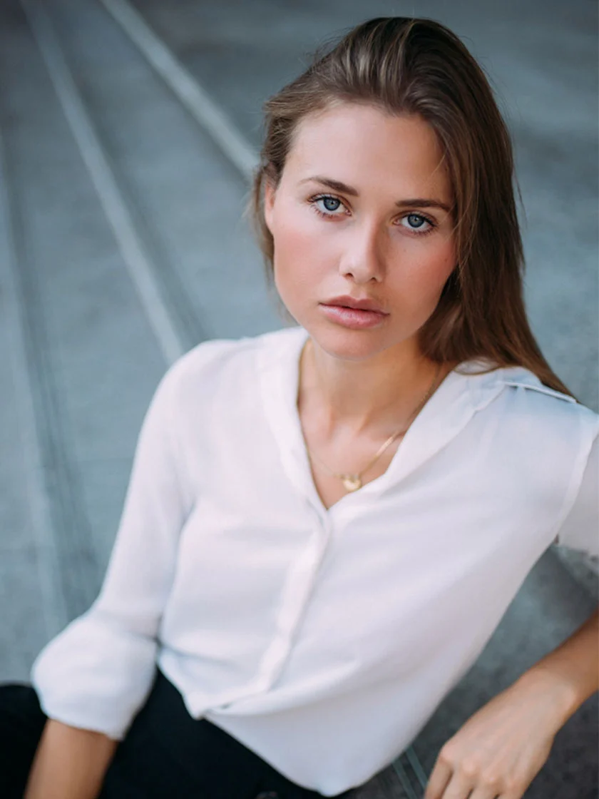 A young woman with long, light brown hair sits facing slightly towards the camera, wearing a white button-down shirt and a gold necklace. Her expression is neutral to serious. The background features a gray-toned surface with faint reflective lines. The lighting is soft and even, highlighting her face and upper body. - Botox in New York and Ridgewood, NJ