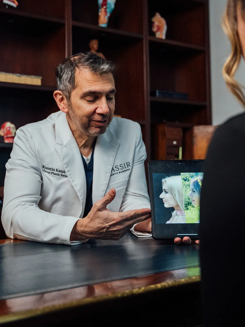 Plastic surgeon Dr. Kassir, in a white coat, consults with a patient at a dark wood desk. He gestures while speaking, and a tablet displays before-and-after facial images. The setting includes dark shelves and soft lighting. - Celebrity Rhinoplasty in New York, NY and Ridgewood, NJ