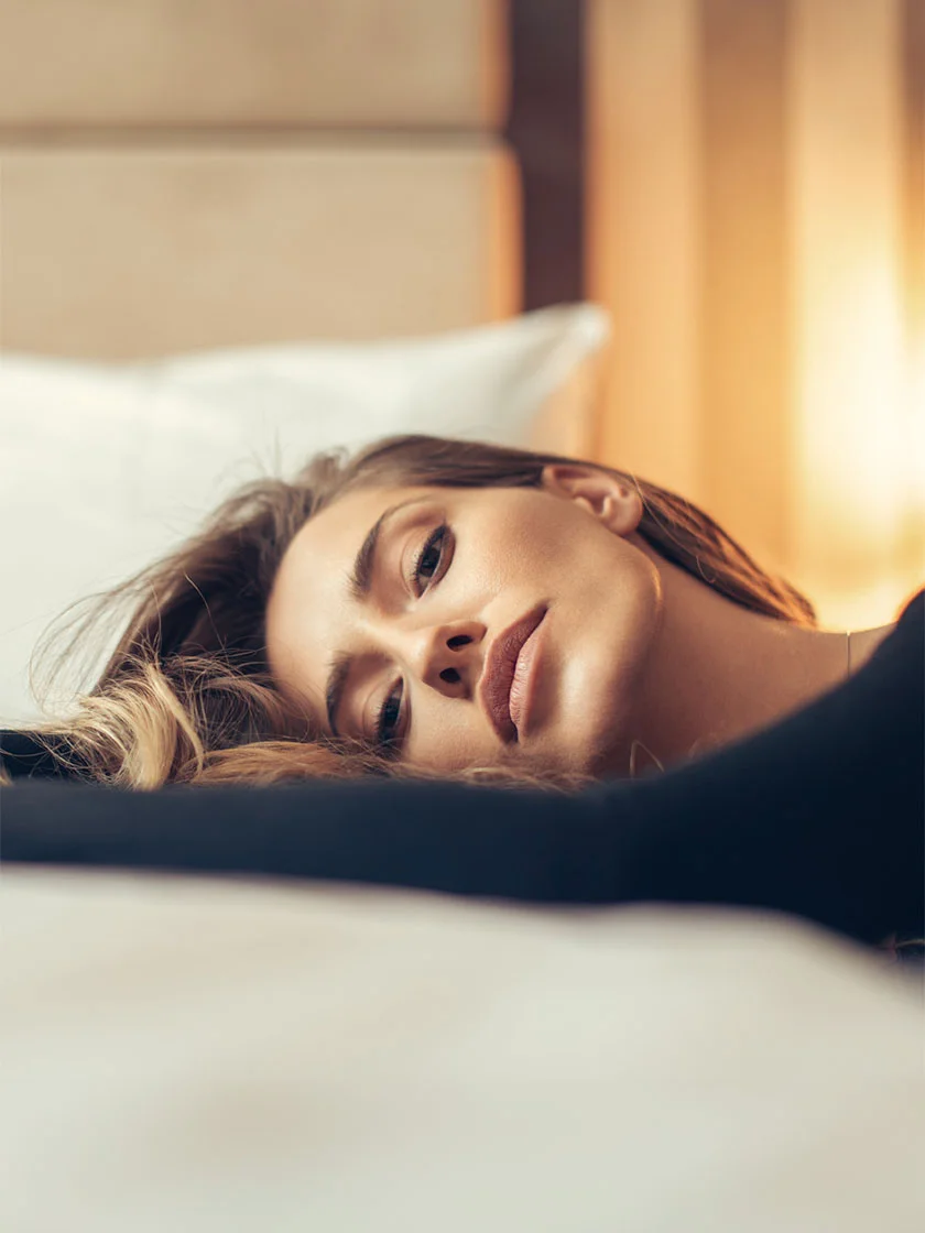 Close-up of a woman lying on a bed with white sheets. She has light brown or blonde hair flowing across her neck and chest, and wears a dark top. Her expression is calm, with a soft gaze directed downward to the left. The background, softly blurred, suggests a bedroom setting in beige tones. - Chin Implant in New York, NY and Ridgewood, NJ