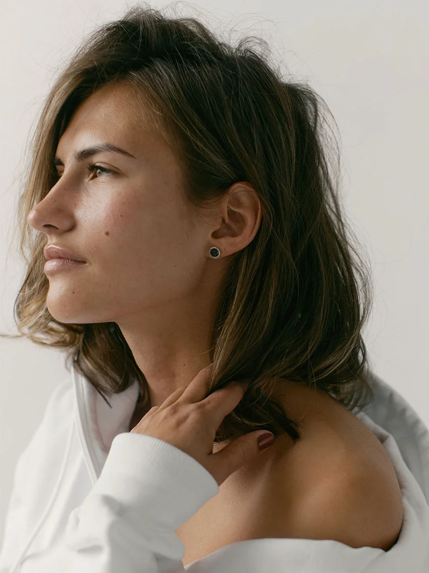 Close-up of a woman with shoulder-length light brown hair, wearing a white top. She has a neutral expression and is looking slightly to the right. Her hand rests gently on her neck near her ear, with a small stud earring visible. The background is a plain, light gray or white wall with soft lighting. - Dermal Fillers in New York, NY and Ridgewood, NJ