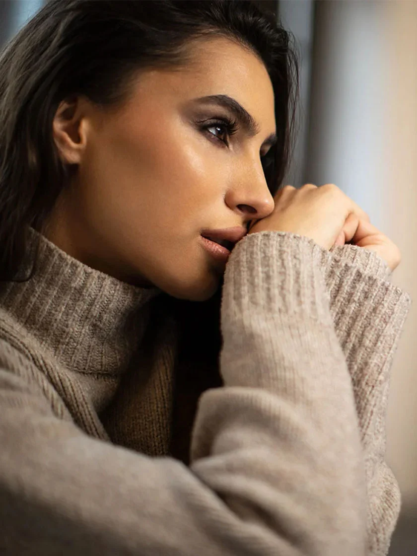 Close-up of a light-skinned woman with long, dark brown hair. She gazes slightly off to the side with a contemplative expression. She wears a light taupe or beige turtleneck sweater with a subtle knit pattern. Her makeup is natural, highlighting her warm brown eyes and muted lips. Her hands gently rest on her face, partially obscuring her cheeks and neck. The background is blurred, creating an intimate portrait feel. - Ethnic Rhinoplasty in New York, NY and Ridgewood, NJ
