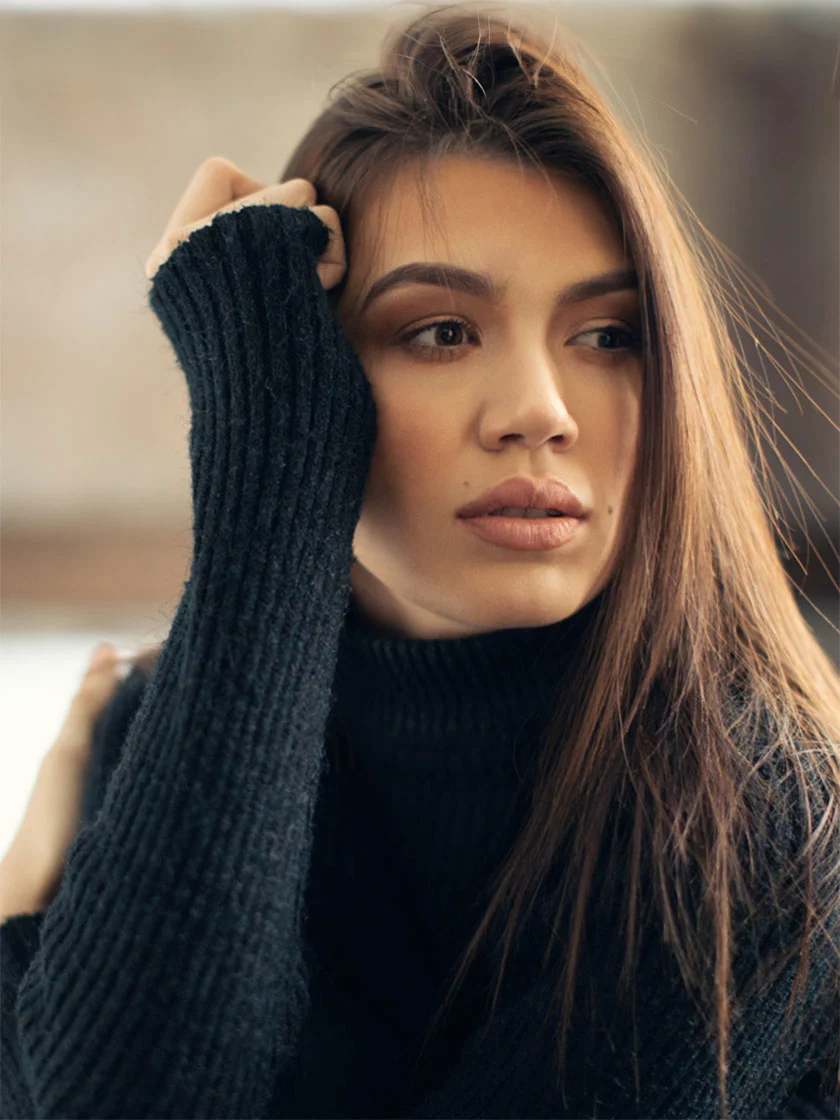 A young woman with light brown hair and a tan complexion is slightly angled towards the camera. Her right arm is raised, gently touching her head. She has a neutral expression, wearing a black one-shoulder tube top and black leggings. The background is light off-white/beige, with soft shadows and highlights, emphasizing the smooth texture of both the background and her skin. - Eyelid Surgery in New York, NY and Ridgewood, NJ
