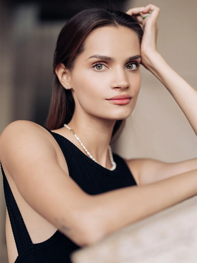 A young woman with shoulder-length dark brown hair, light-beige skin, and green eyes gazes neutrally. She wears a black ribbed sleeveless top, a pearl necklace, and stud earrings. Her right arm is bent and laid on her head, while her left arm rests relaxed. The blurred background is a light beige, suggesting an indoor setting. - Fat Transfer in New York, NY and Ridgewood, NJ