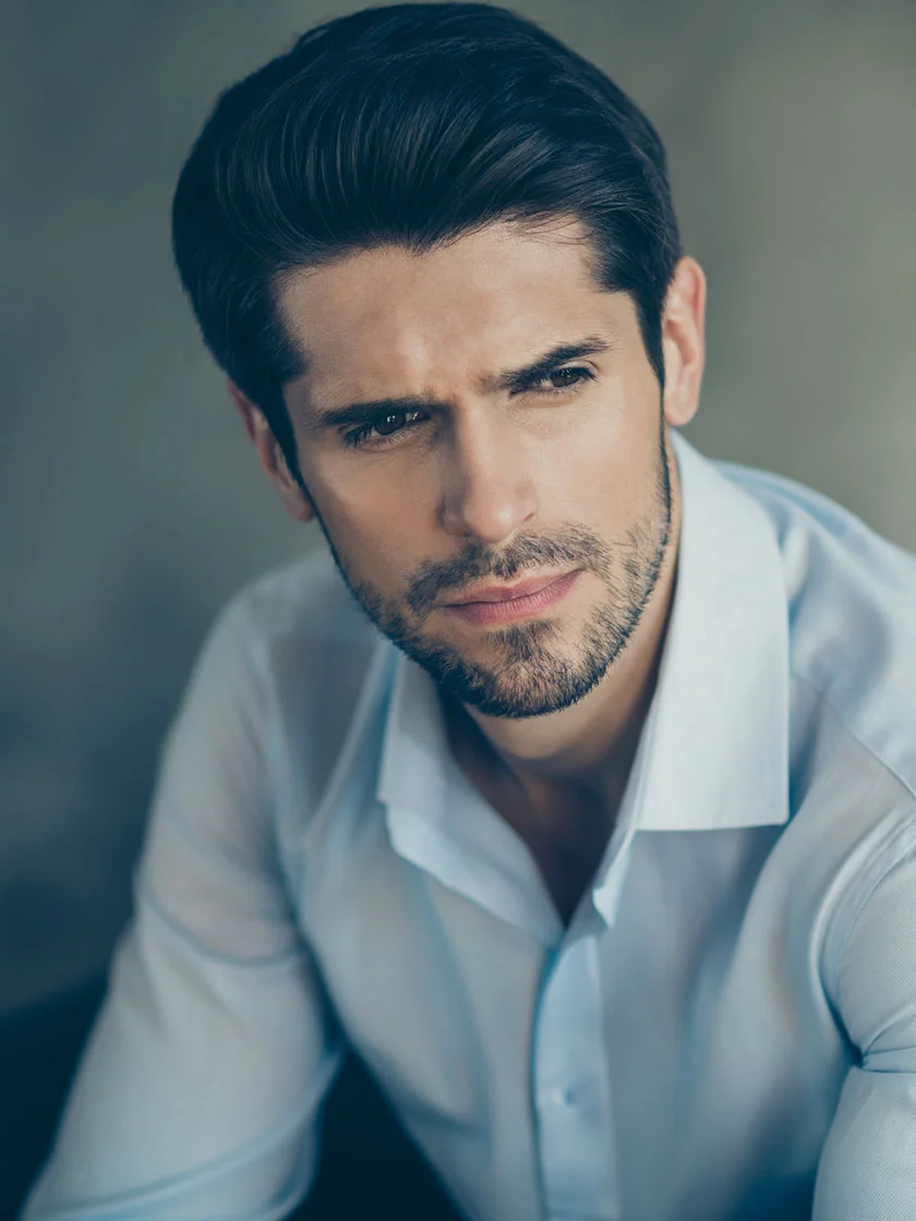 Close-up of a man with short, dark hair styled back and a neatly trimmed dark brown beard. He wears a light blue or pale gray dress shirt with a button-down collar. His expression is contemplative, and his dark eyes and brows are sharply defined. The background is a muted gray or taupe, with even lighting emphasizing his face and upper torso. - Male Rhinoplasty in New York, NY and Ridgewood, NJ