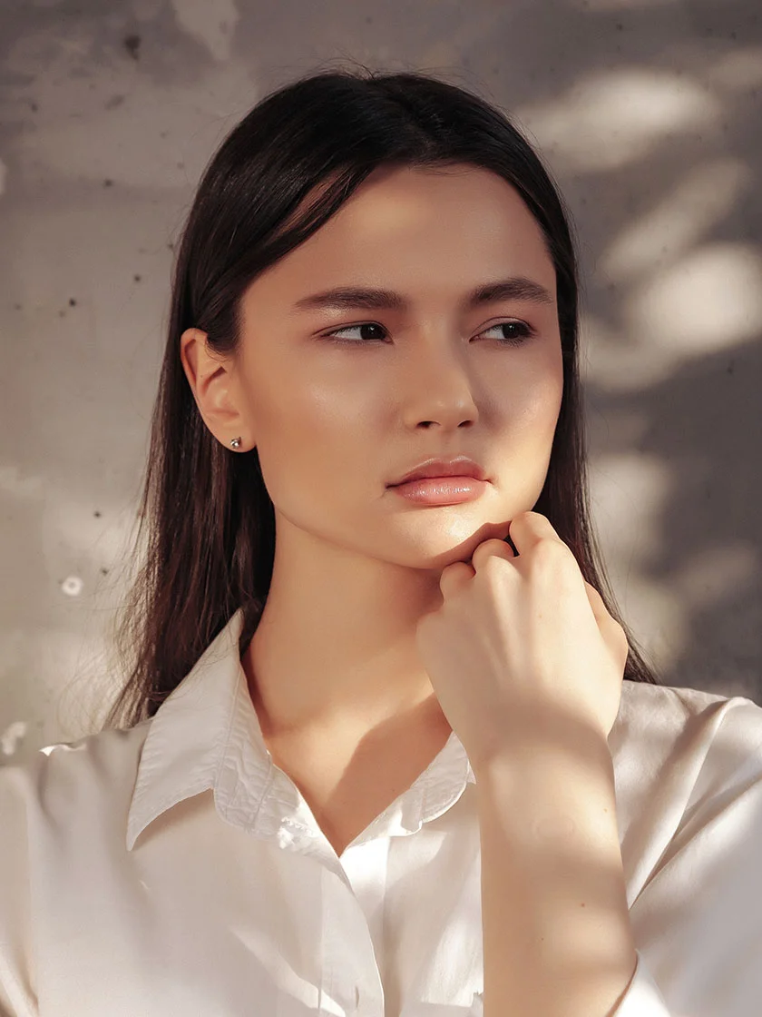 Close-up of a young woman with shoulder-length dark hair, wearing a white button-up shirt. Her light-toned skin is softly lit, and her expression is thoughtful as she gazes off to the side. One hand rests near her chin. The background is a textured, muted concrete wall with gentle shadows. - Microneedling in New York, NY and Ridgewood, NJ