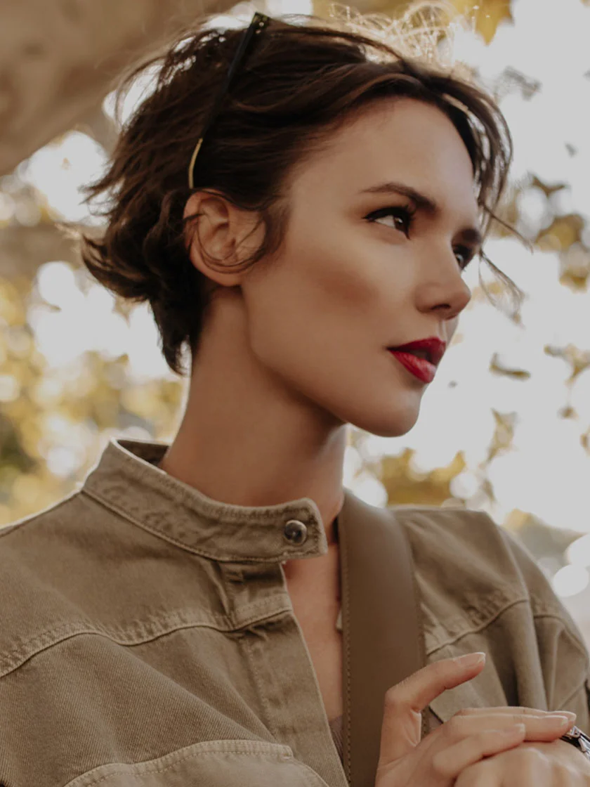 Close-up portrait of a young woman with short, wavy brown hair, wearing a thin gold headband. She has vibrant red lips and a neutral, thoughtful expression while looking to the right. She is dressed in a light brown, collared, button-up jacket. The softly lit background is outdoors and out of focus, featuring warm-toned natural elements like trees or foliage. - Non-Surgical Rhinoplasty in New York, NY and Ridgewood, NJ