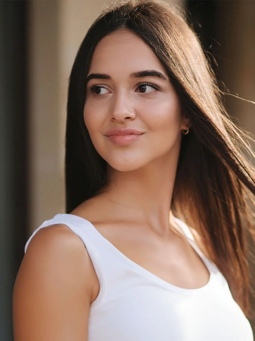 Close-up of a young woman with long, dark brown hair, wearing a white sleeveless top. She has a neutral expression, with a soft smile and light-toned, smooth skin. The background is blurred, suggesting an outdoor setting with natural light highlighting her features. - Teen Rhinoplasty in New York, NY and Ridgewood, NJ