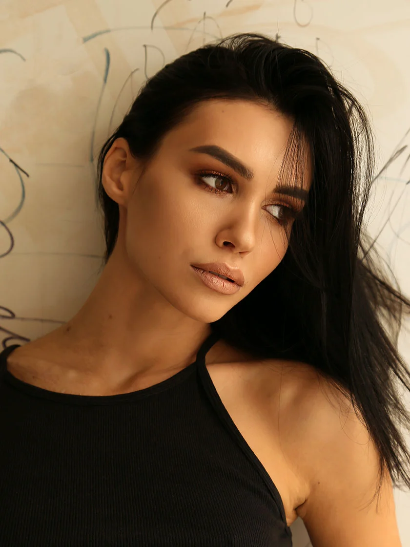 A woman with long, dark, slightly tousled hair wears a black ribbed halter top. Her light brown skin glows with natural makeup, including subtle neutral eyeshadow and a soft lip color. Her hazel-brown eyes and defined eyebrows enhance her relaxed expression as she slightly turns toward the viewer’s left. The background features a light beige or cream-colored wall with faint markings, adding a soft, artistic touch. - Thread Lifts in New York, NY and Ridgewood, NJ